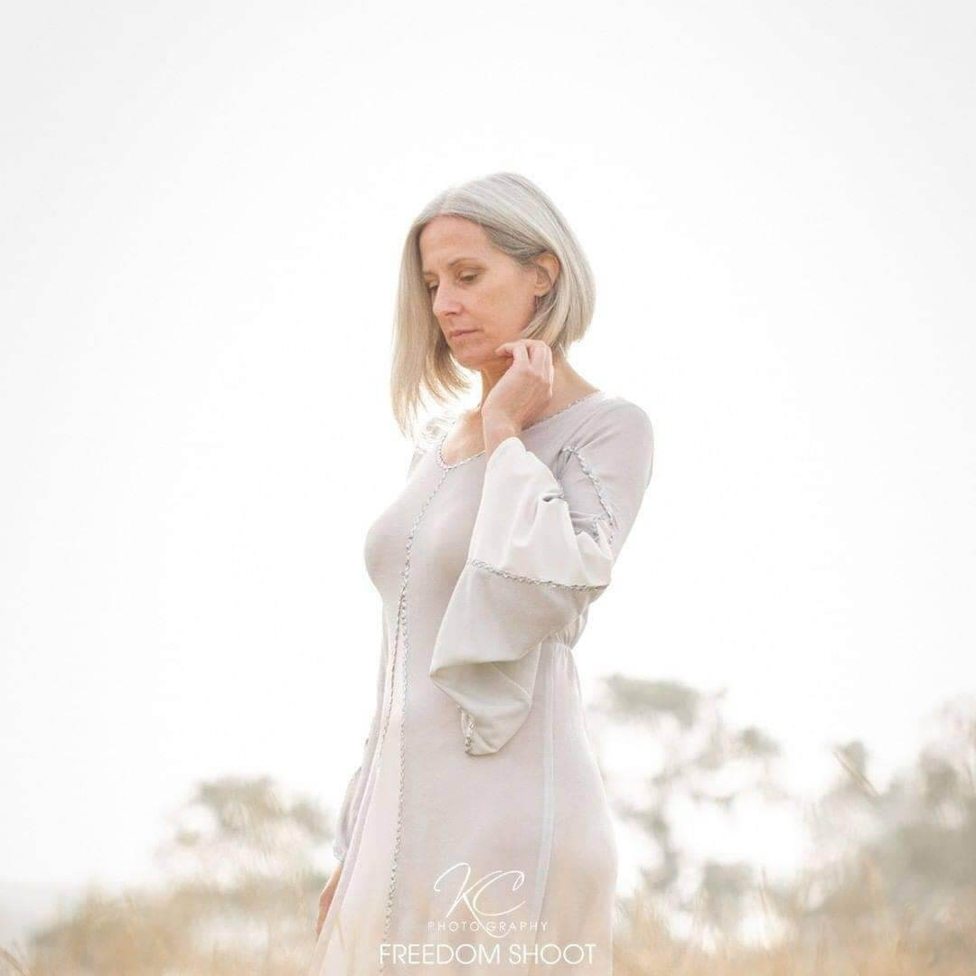 White gentle and soft photograph outside of a lady in a long white dress and short white hair and hand up to chin